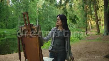 一位穿着衬衫和牛仔裤的漂亮女艺术家在池塘附近的公园里画一幅风景画
